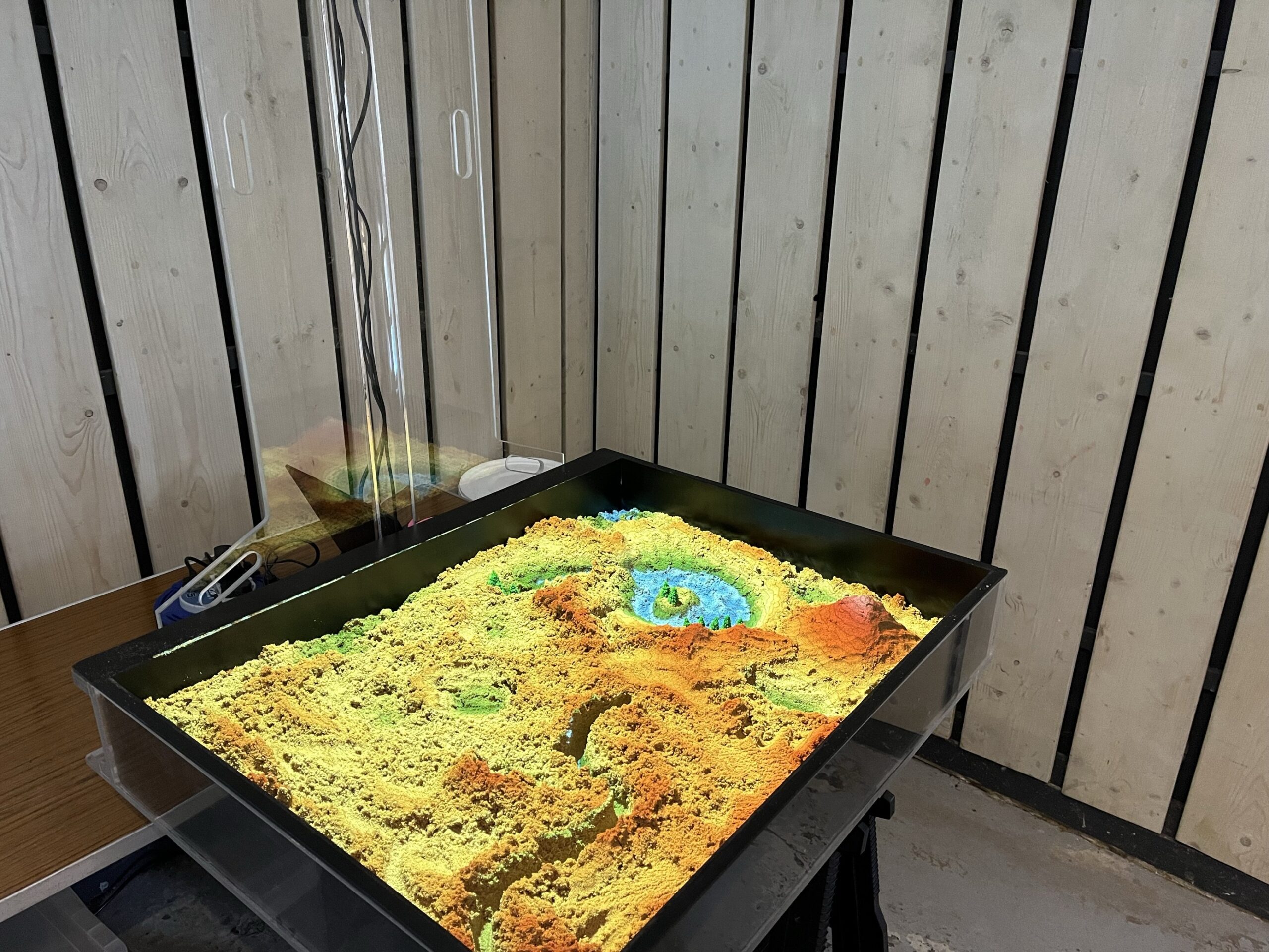 Image shows an AR Sandbox fully set up. The image has been taken from one of the corners of the sandbox and shows the sand lit up by the projector with different hills, slopes and a pond.